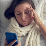 Kobieta leży w łóżku, sprawdza w telefonie objawy koronawirusa.