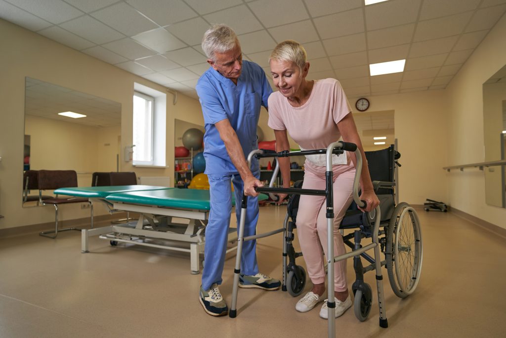 Pacjentka podczas rehabilitacji neurologicznej wstaje z wózka i przy pomocy balkoniku oraz wsparciu fizjoterapeuty uczy się chodzić