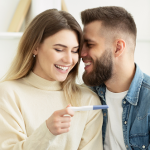 Uśmiechnięta kobieta w 4. tygodniu ciąży trzyma test ciążowy, obok siedzi uśmiechnięty mężczyzna
