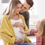Uśmiechnięta kobiet w 5. miesiącu ciąży siedzi na ziemi z córką i mężem - dziecko trzyma rękę na brzuchu ciążowym