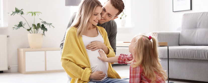 Uśmiechnięta kobiet w 5. miesiącu ciąży siedzi na ziemi z córką i mężem - dziecko trzyma rękę na brzuchu ciążowym